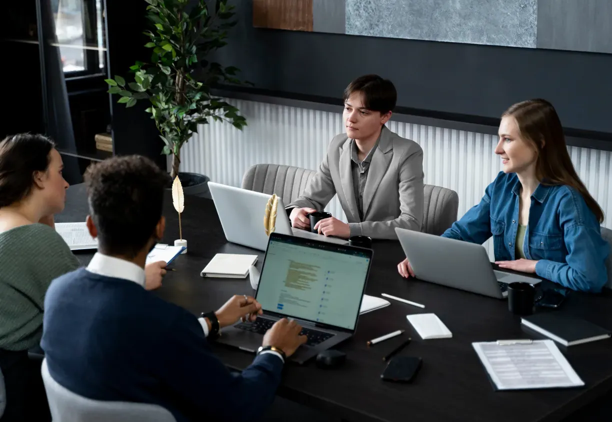 Four colleagues in a meeting around a table with open laptops and notebooks