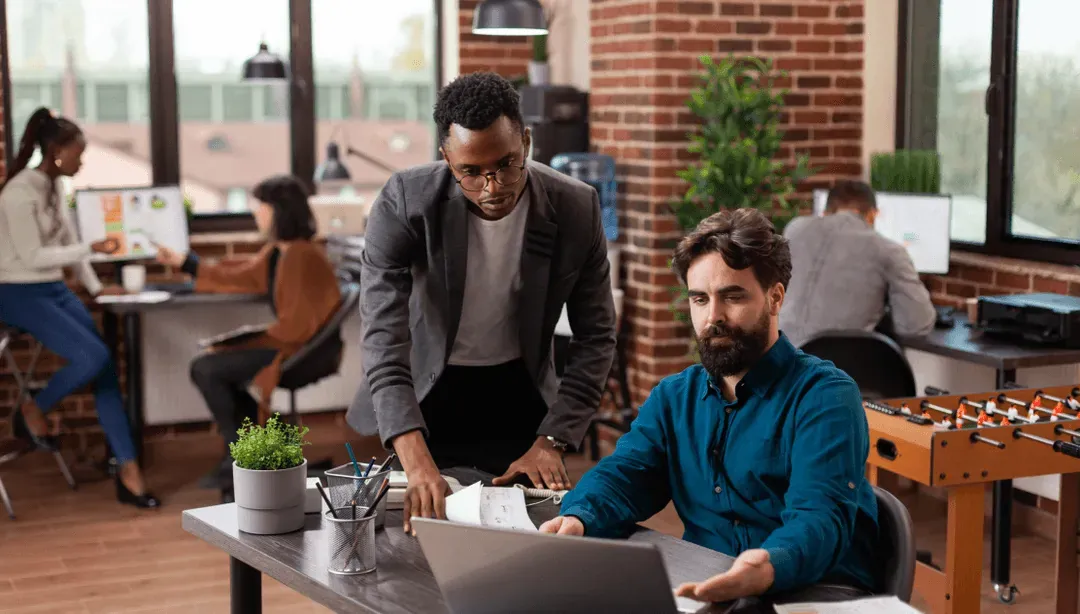 Two colleagues reviewing work on a laptop in a brick-walled office