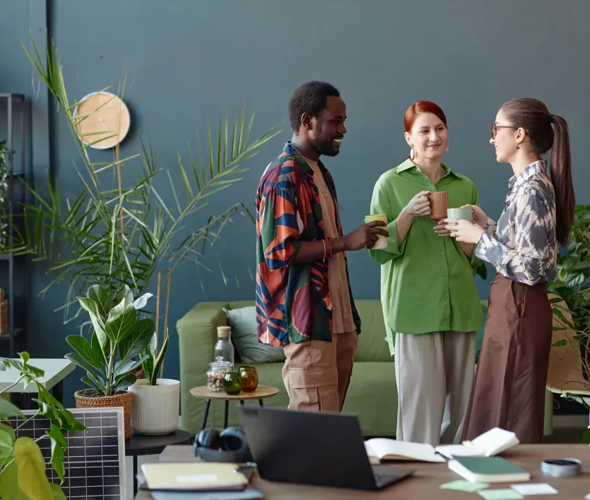 Three colleagues chatting over coffee in a studio with plants and a desk of notebooks